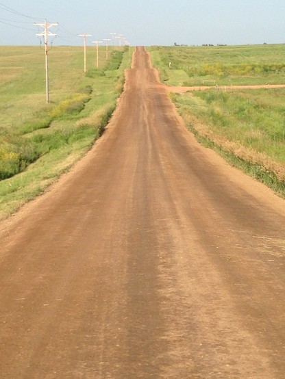Looking down a brown dirt road which heads over low hills towards the horizon and blue sky. Green grass on either side, and an electrical line on the left side.