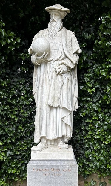 White statue of a man with a long beard, wearing robes and a floppy hat, nestling a globe in his right arm and holding a protractor in his left hand